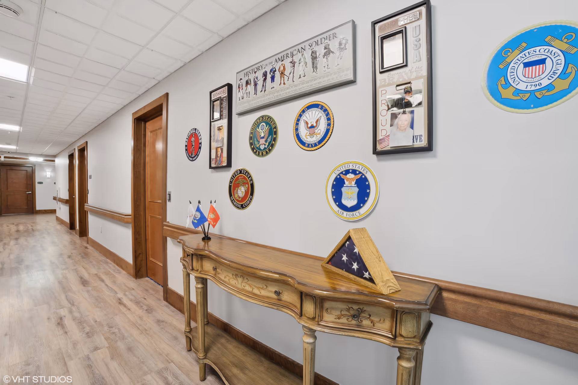 A hallway in a senior living facility with wooden doors and a wooden console table against the wall. Above the table are various military emblems and plaques, including those of the United States Army, Navy, Marine Corps, Air Force, and Coast Guard. The wall also features a framed display titled 'History of the American Soldier' and small flags on the table. The floor is wood, and the ceiling has white tiles with fluorescent lighting.