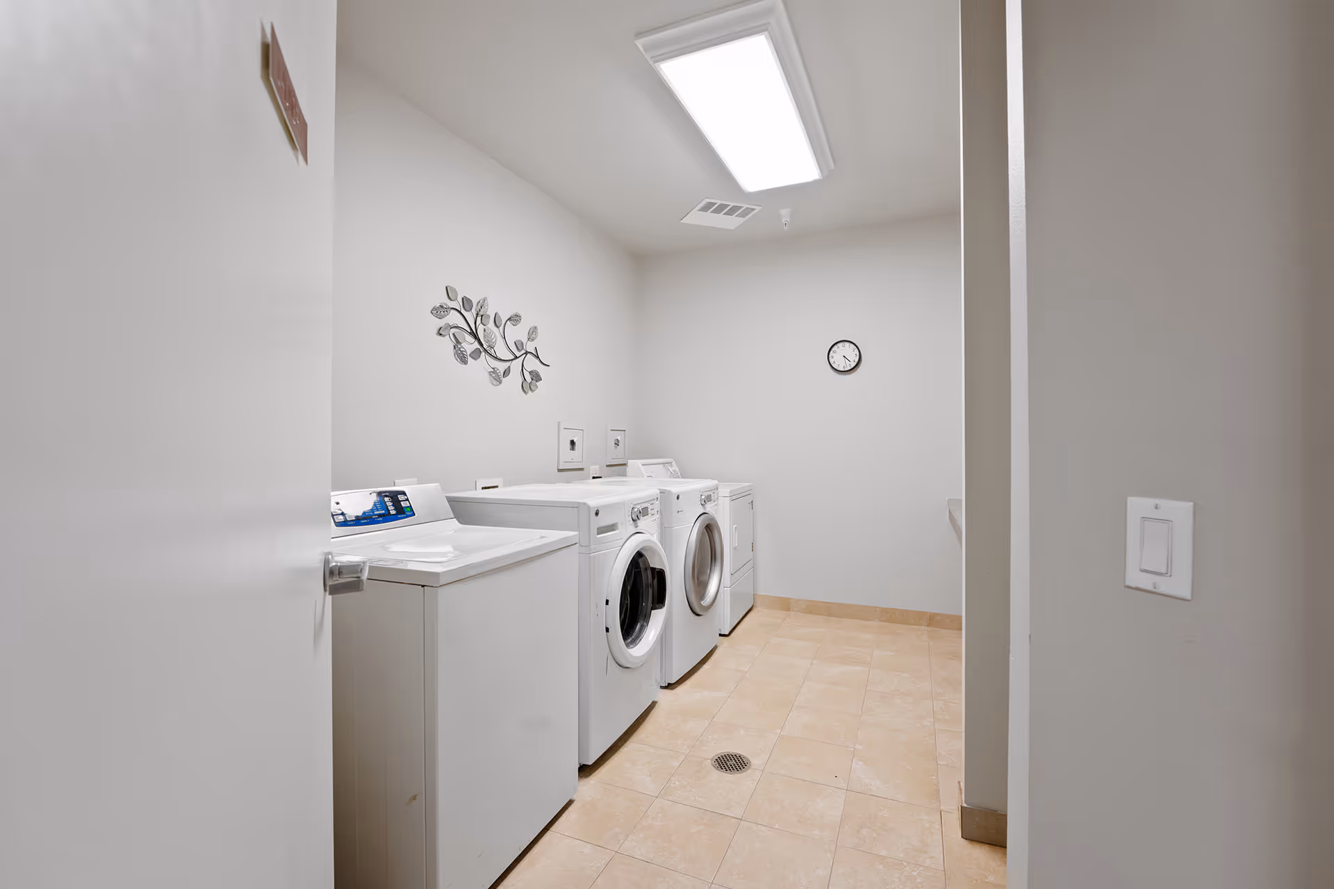 A clean laundry room with front-loading washer and dryer units along a pale wall, tile floor, wall clock, and overhead light.