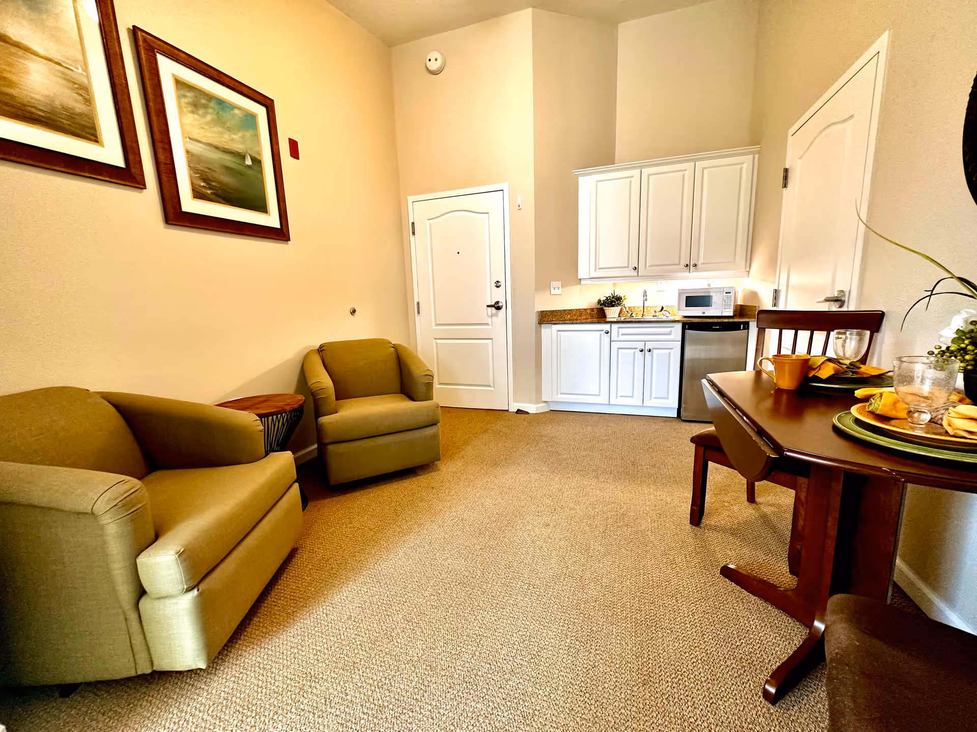 Interior view of a senior living facility room featuring two beige armchairs, a small round wooden side table, a kitchenette with white cabinets, a microwave, and a mini fridge. A wooden dining table set for two with yellow napkins and glassware is visible on the right. The walls are decorated with framed paintings, and the room has beige carpeting.