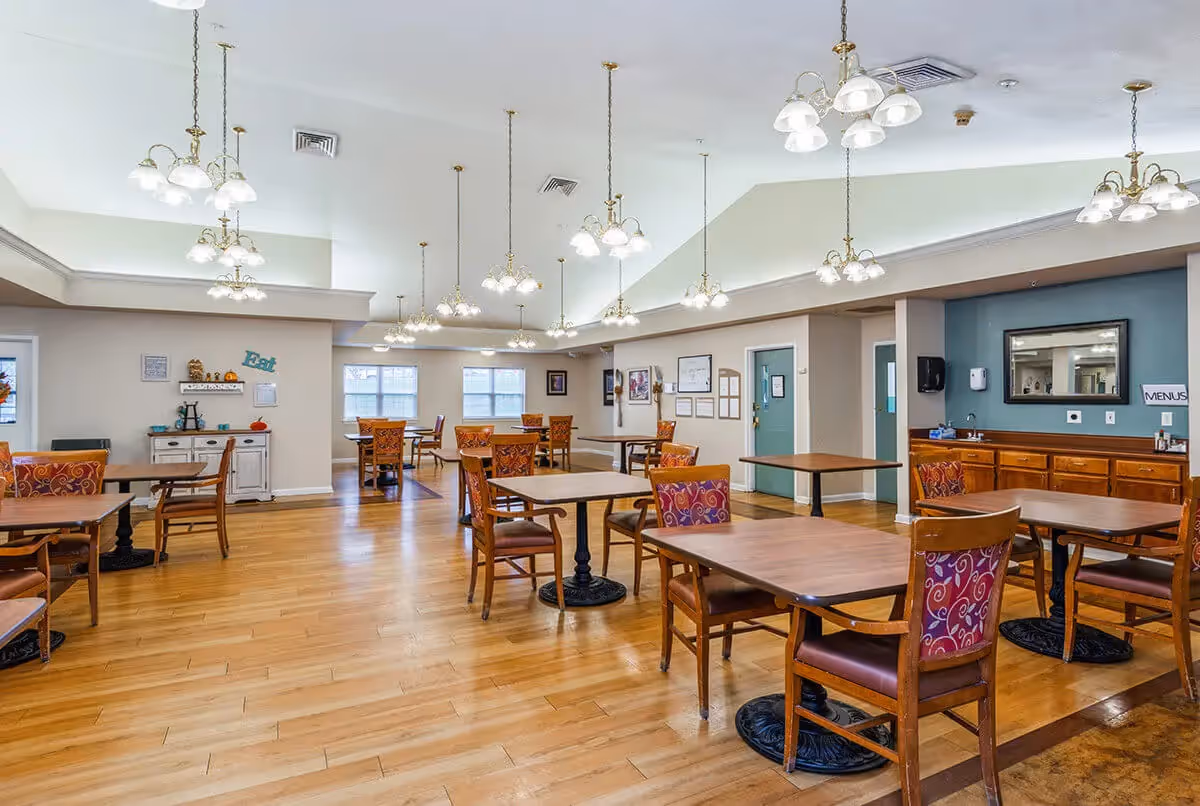 A spacious dining room with wooden floors and multiple wooden tables and chairs with patterned upholstery. The room is well-lit with several hanging light fixtures from a high ceiling. There is a sideboard with a mirror and a sign that says 'MENUS' on the right wall, and a small white cabinet with decorative items and a sign that says 'Eat' on the left wall. The room has large windows letting in natural light.