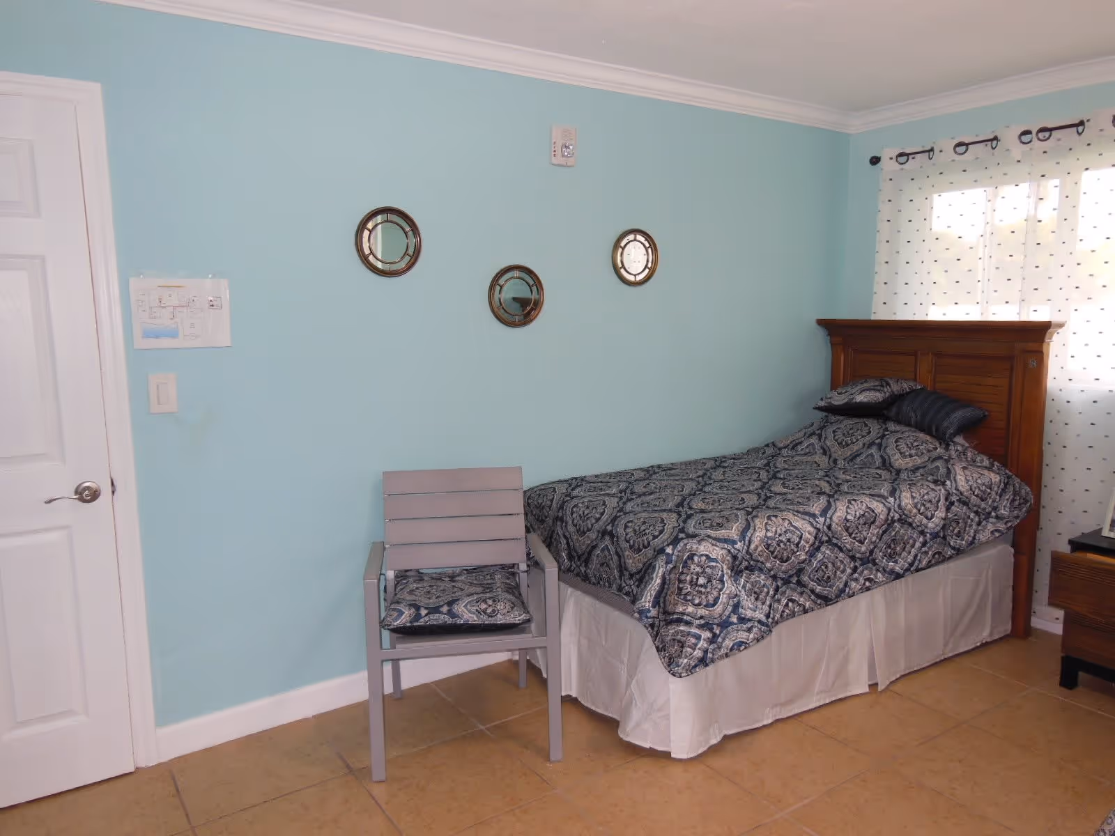 A bedroom with a single bed featuring a wooden headboard and patterned blue and white bedding. Next to the bed is a gray chair with a matching cushion. The walls are painted light blue with three round decorative mirrors hanging above the chair. A window with white curtains with small blue dots is on the right side, and a white door is on the left side of the room.