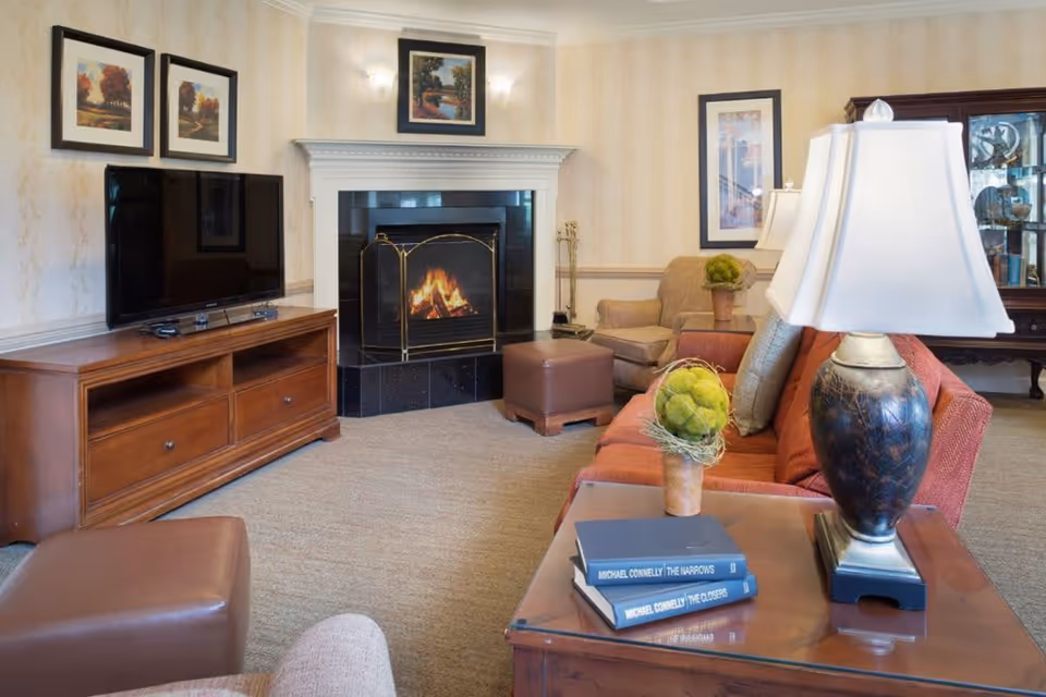 A cozy living room with a lit fireplace, a flat-screen TV on a wooden stand, a red sofa with cushions, a brown armchair, a brown ottoman, a wooden side table with a lamp and decorative green artichokes, and framed artwork on the walls.