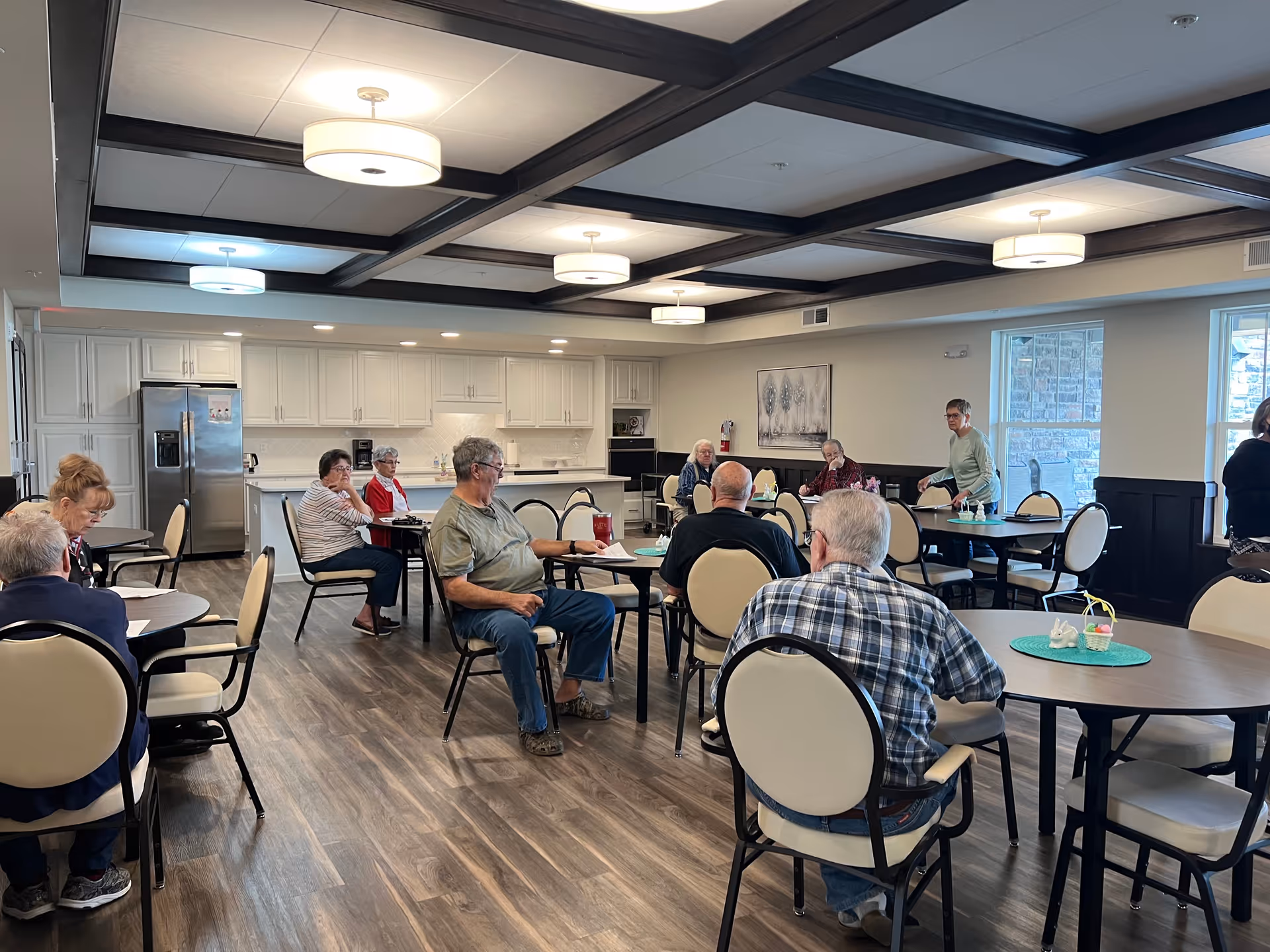 Seniors seated at round tables in a bright communal dining/activity room with a kitchen area in the background.