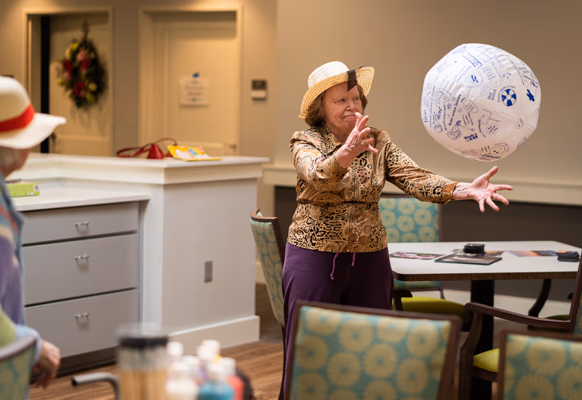An elderly woman wearing a hat and patterned jacket is playing with a large inflatable ball indoors, while another person wearing a hat watches. The room has chairs with patterned upholstery, a table with magazines, and a counter with some items on it.