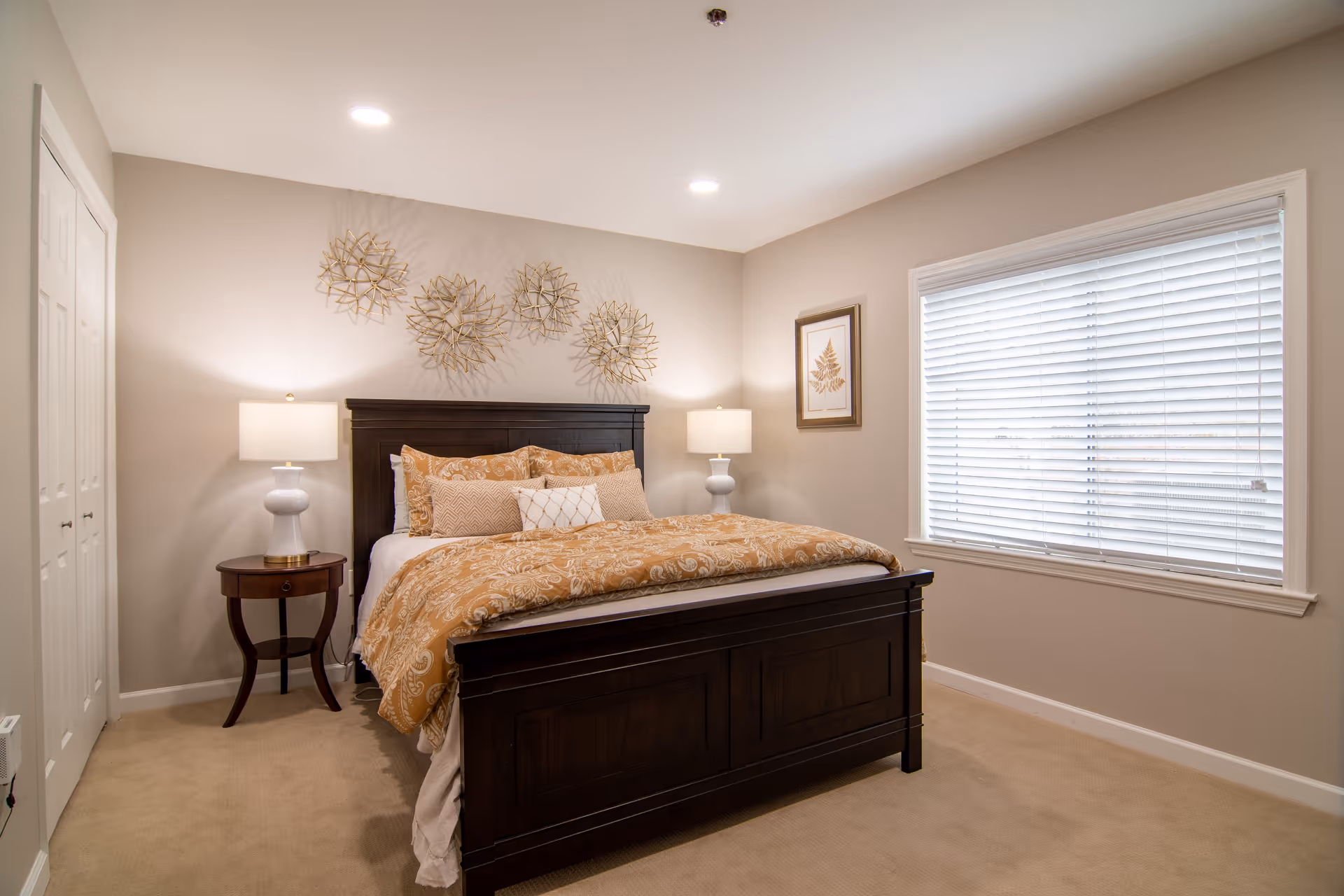 A cozy bedroom in a retirement community featuring a dark wooden bed frame with a beige and gold patterned comforter and multiple pillows. There are two white lamps on either side of the bed on small wooden nightstands. The walls are painted light beige with decorative metal wall art above the headboard and a framed botanical print on the adjacent wall. A large window with white blinds allows natural light into the room.