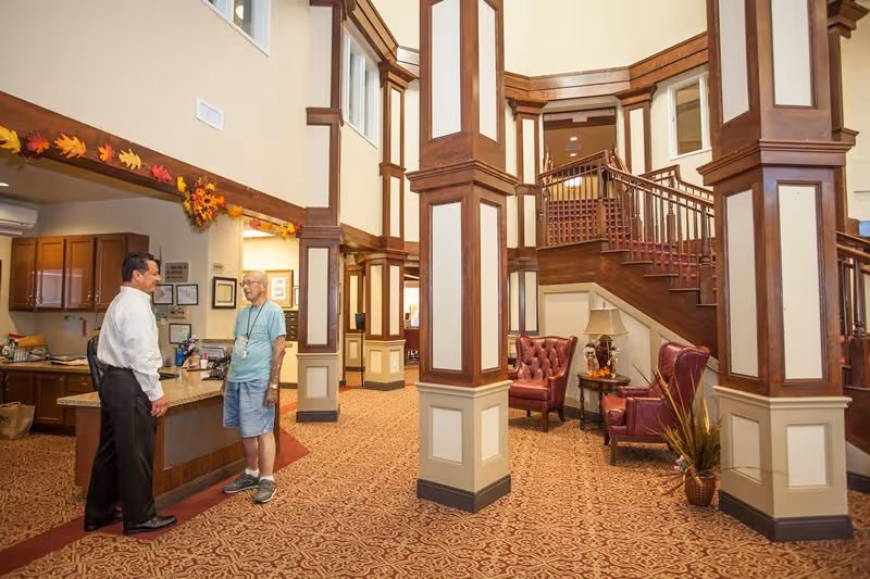 Spacious assisted living lobby with wood-paneled columns and staircase, a reception desk, seating area, and two people talking.