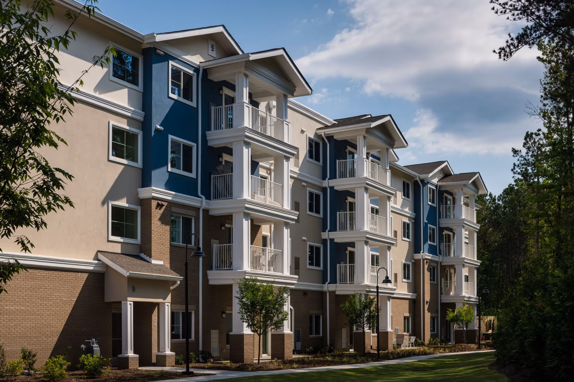 Exterior view of a multi-story residential building with beige and blue walls, white balconies, and a well-maintained lawn and trees in front under a partly cloudy sky.