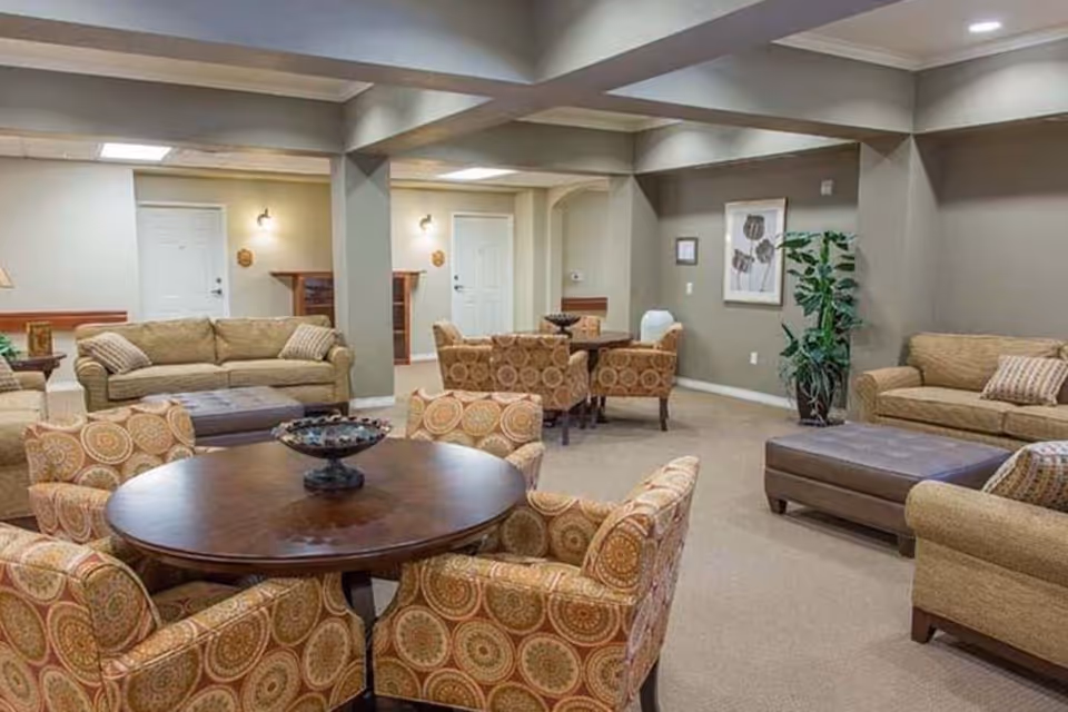 A spacious living room area in a senior living facility featuring multiple beige sofas, patterned armchairs around a round wooden table, a large ottoman, a potted plant, and framed artwork on the wall. The room has neutral-colored walls and carpeted floors with ceiling beams and recessed lighting.