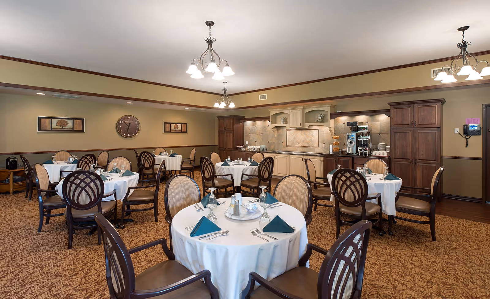 Dining room with round tables draped in white tablecloths, green napkins, wooden chairs, and a serving counter/kitchen area along the back wall.