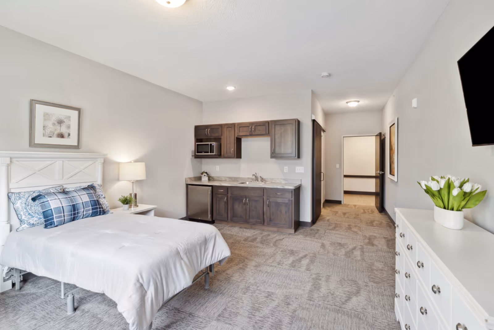 A senior living facility room featuring a bed with white bedding and blue patterned pillows, a nightstand with a lamp and small plant, a kitchenette with dark wood cabinets, a microwave, sink, and mini fridge, a white dresser with a vase of white tulips, a wall-mounted TV, and a hallway leading to another room.
