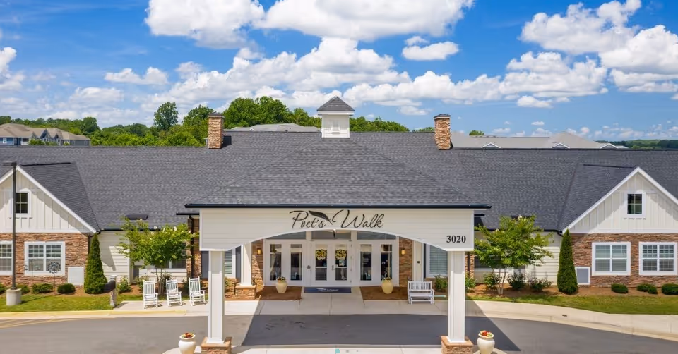 Front exterior view of Poet's Walk Warrenton, A Memory Care Community, showing a large building with a covered entrance, white pillars, and a sign with the facility's name. The building has a dark shingled roof, brick and white siding walls, and is surrounded by greenery under a partly cloudy blue sky.