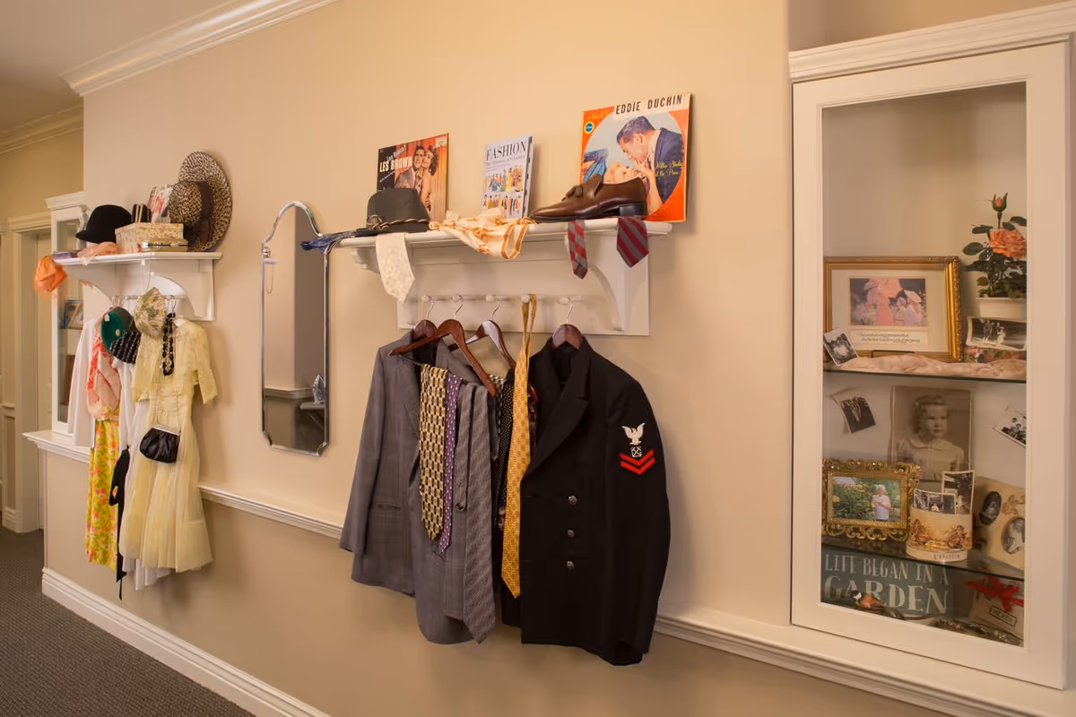 A hallway wall display featuring vintage clothing including hats, dresses, jackets, and ties hanging on white shelves and hooks. There are also vintage magazines and shoes placed on the shelves above the clothing. To the right, a glass cabinet contains framed photographs and decorative items.