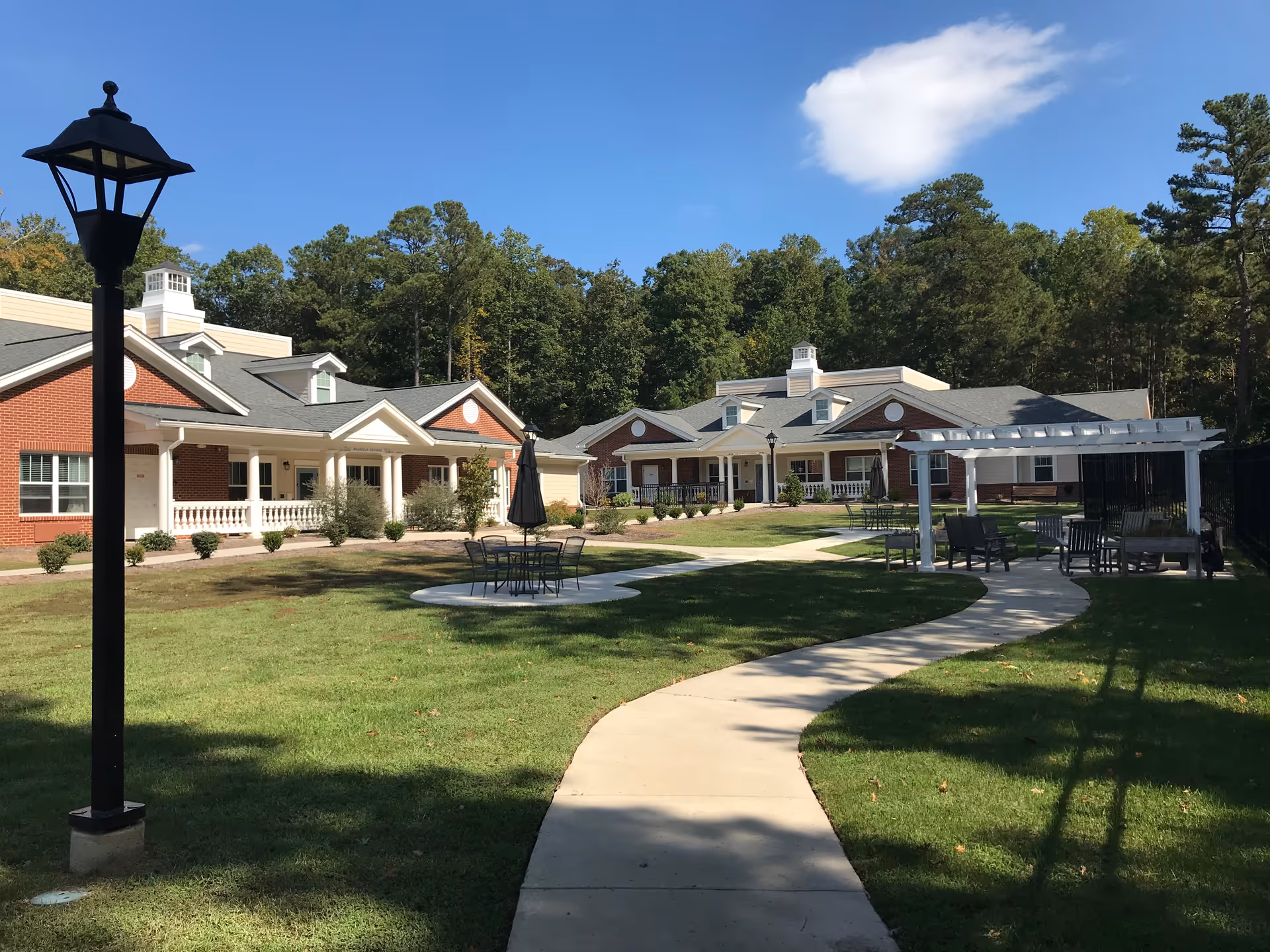 Outdoor view of English Meadows Williamsburg Campus showing a paved walkway curving through a grassy courtyard with outdoor seating areas, lamp posts, and red brick buildings with white trim surrounded by trees under a blue sky with a few clouds.