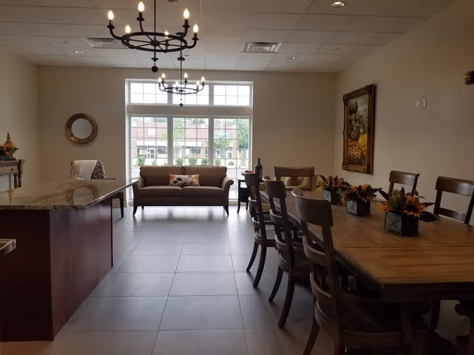 A spacious and well-lit interior room featuring a large wooden dining table with eight chairs, decorated with small flower arrangements. To the left, there is a kitchen island with a granite countertop and a chair. At the back, a brown sofa is positioned in front of large windows letting in natural light. Two black chandeliers hang from the ceiling, and a framed painting is on the right wall.
