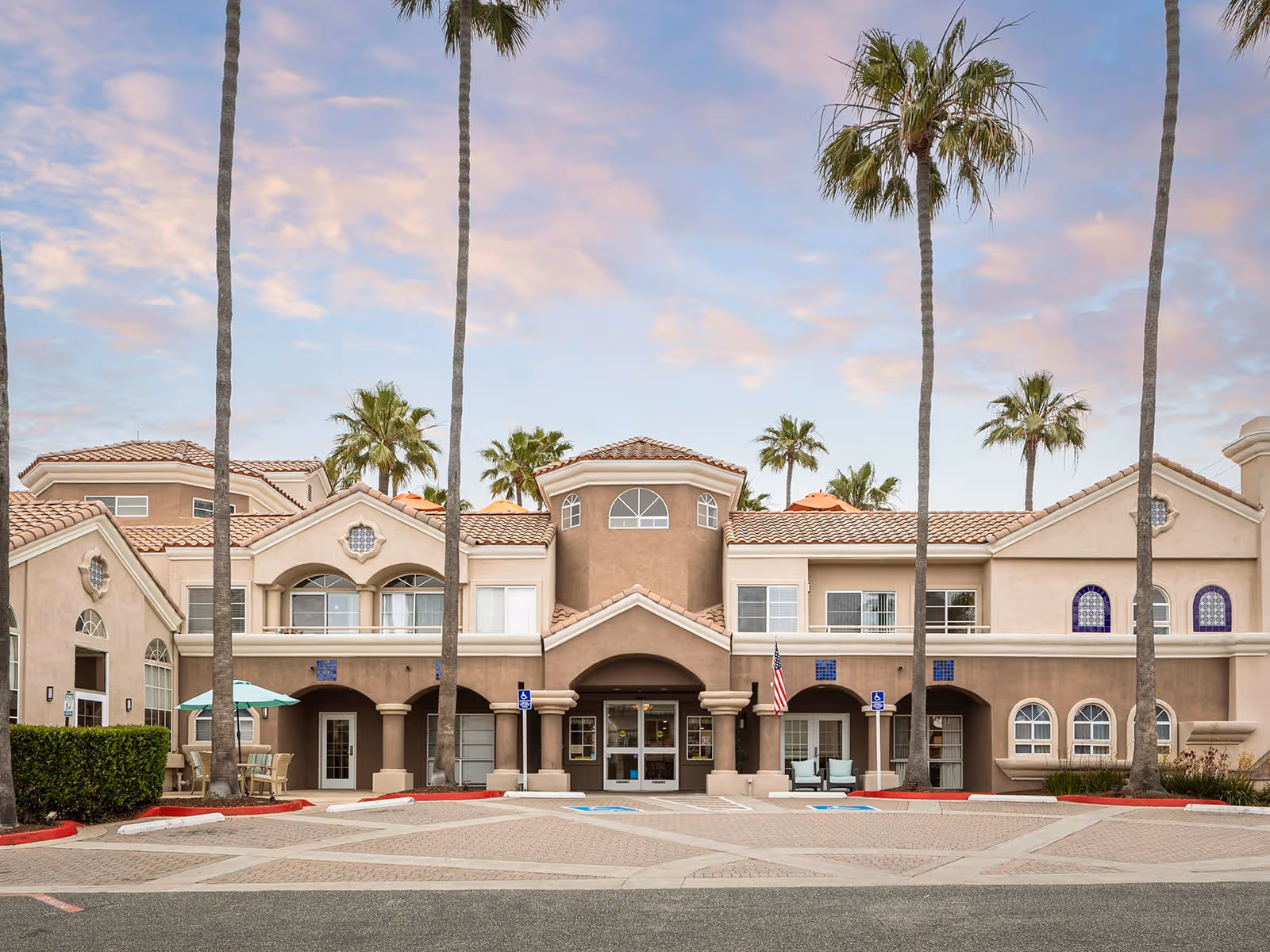 Front exterior view of Atria Newport Plaza, a two-story building with a beige and brown stucco facade, arched windows, and a tiled roof. Tall palm trees line the entrance area, which includes a covered entryway with columns, outdoor seating, and handicap parking spaces in front.