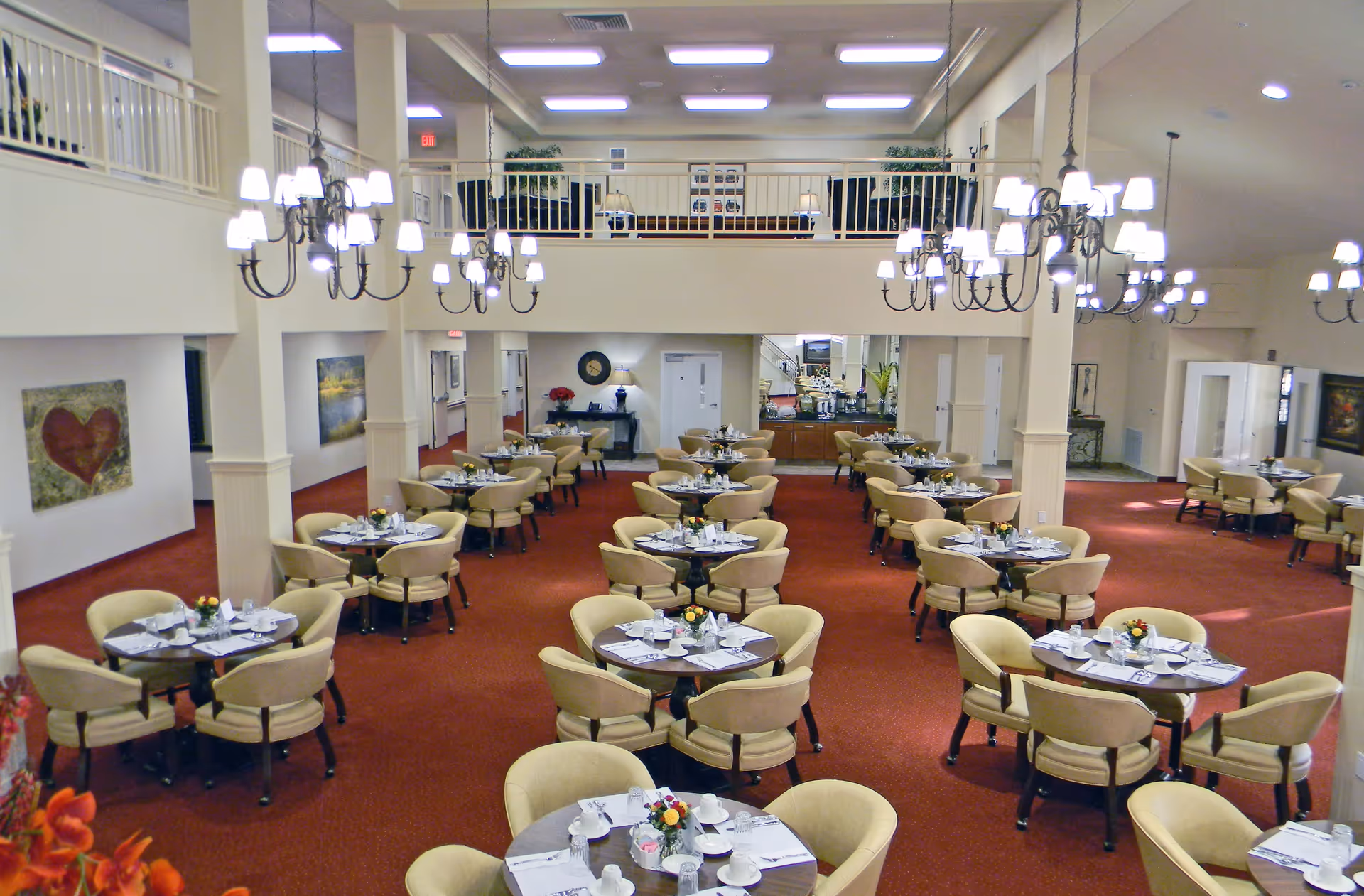 Spacious dining room with multiple round tables set with white cups, plates, and silverware. Each table has four beige upholstered chairs and a small floral centerpiece. The room features red carpet, cream-colored walls, several chandeliers hanging from the ceiling, and artwork on the walls. There is a balcony area above and a buffet or serving area at the back.