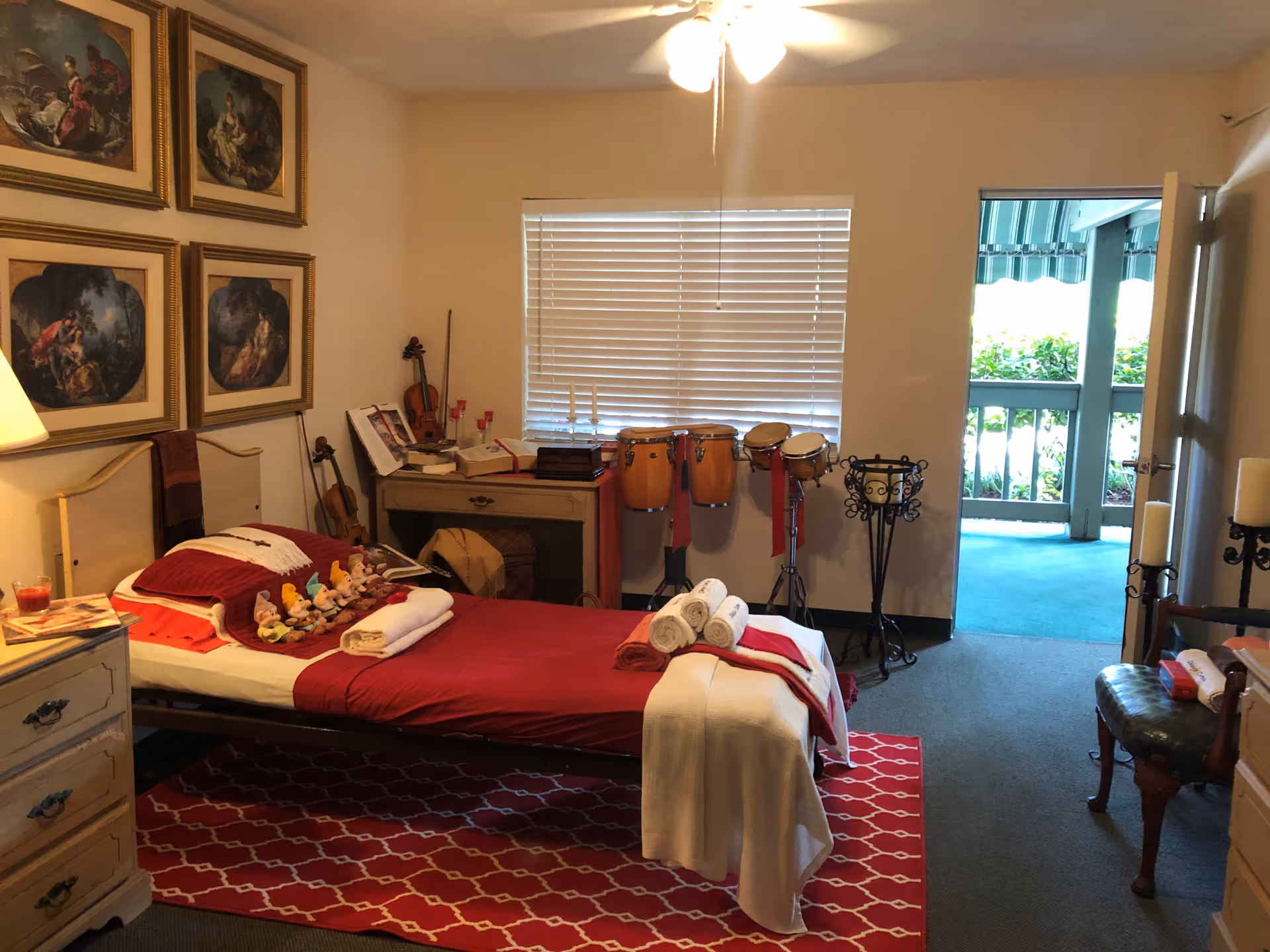 A cozy bedroom with a single bed dressed in red linens, framed paintings on the wall, musical instruments by a desk, and a door leading to an outdoor walkway.