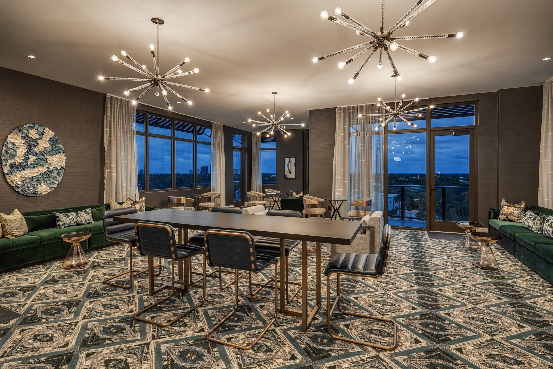 A spacious, modern common area with large windows showing an evening cityscape. The room features a long table with six black chairs, two green velvet sofas with decorative pillows, small round side tables, and patterned carpet. Three contemporary starburst chandeliers hang from the ceiling, and sheer curtains frame the windows and glass door leading to a balcony.