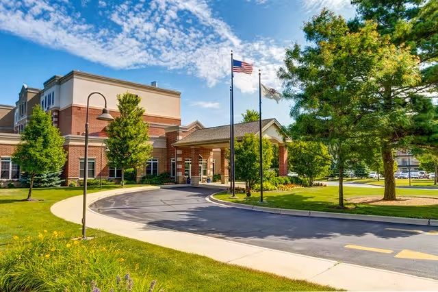 Exterior view of Montclair Assisted Living and Memory Care facility showing a curved driveway leading to the entrance, with two flagpoles flying the American flag and another flag. The building is a multi-story brick structure surrounded by green trees and well-maintained landscaping under a partly cloudy blue sky.