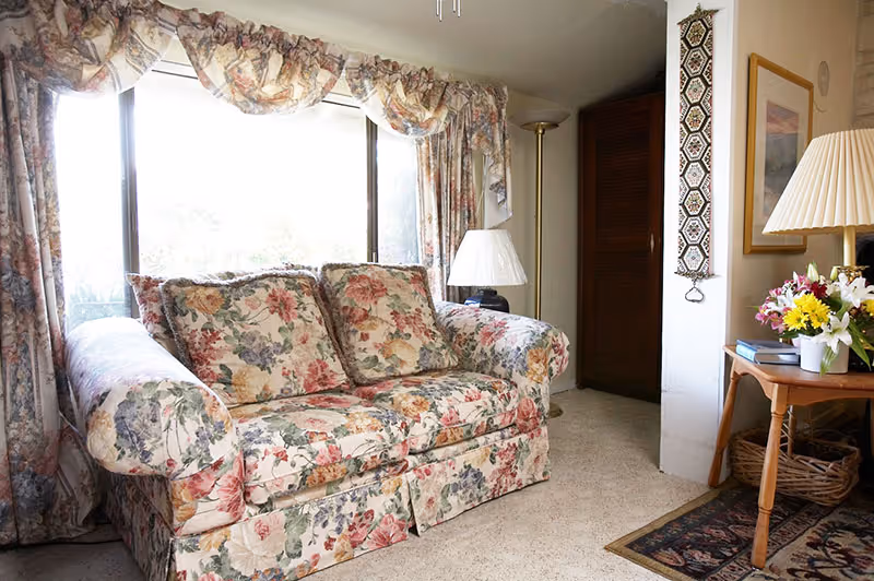 Cozy living room with a floral-patterned loveseat in front of a large window, flanked by lamps and a side table with a vase of flowers.