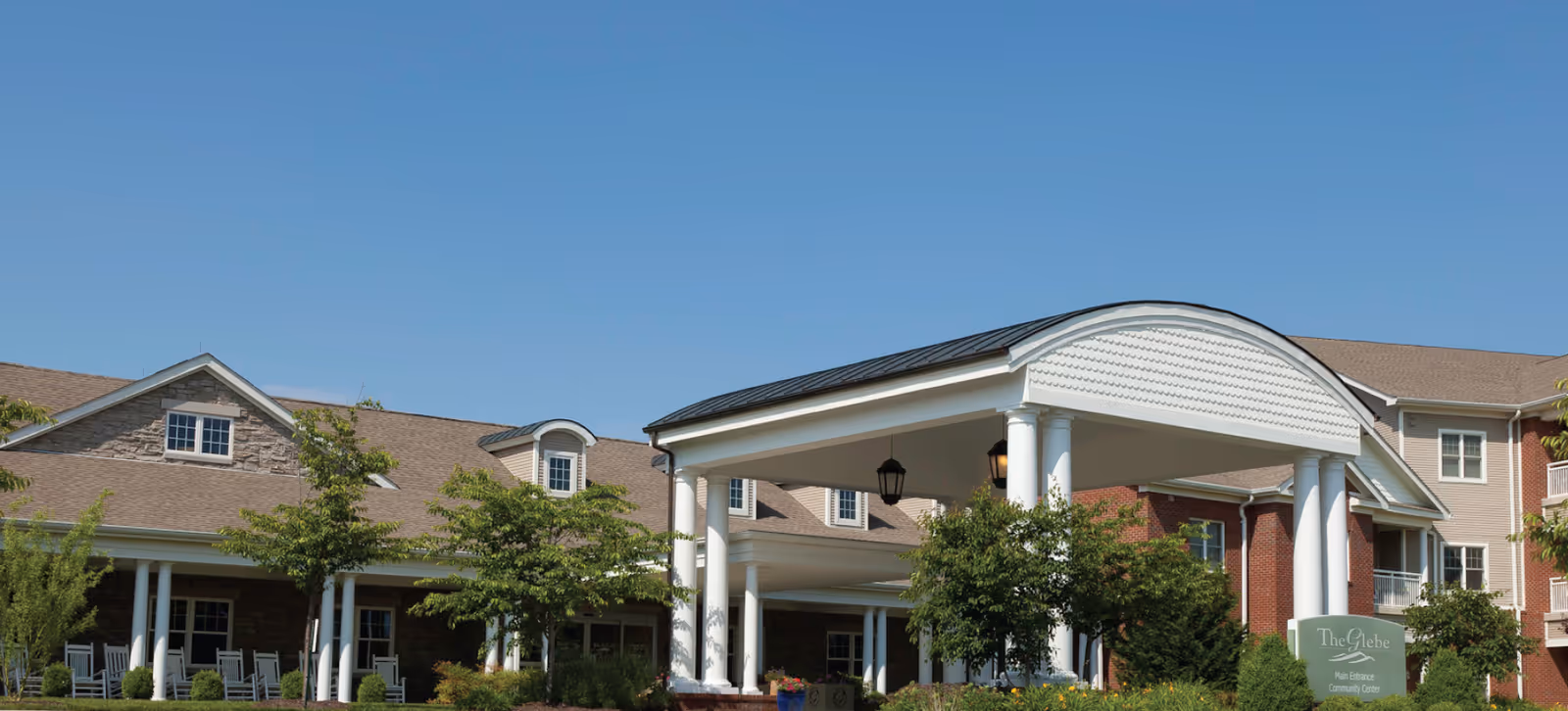 Front entrance of The Glebe Retirement Community showing a covered porte-cochère with white columns, brick building, and landscaped grounds under a clear blue sky.