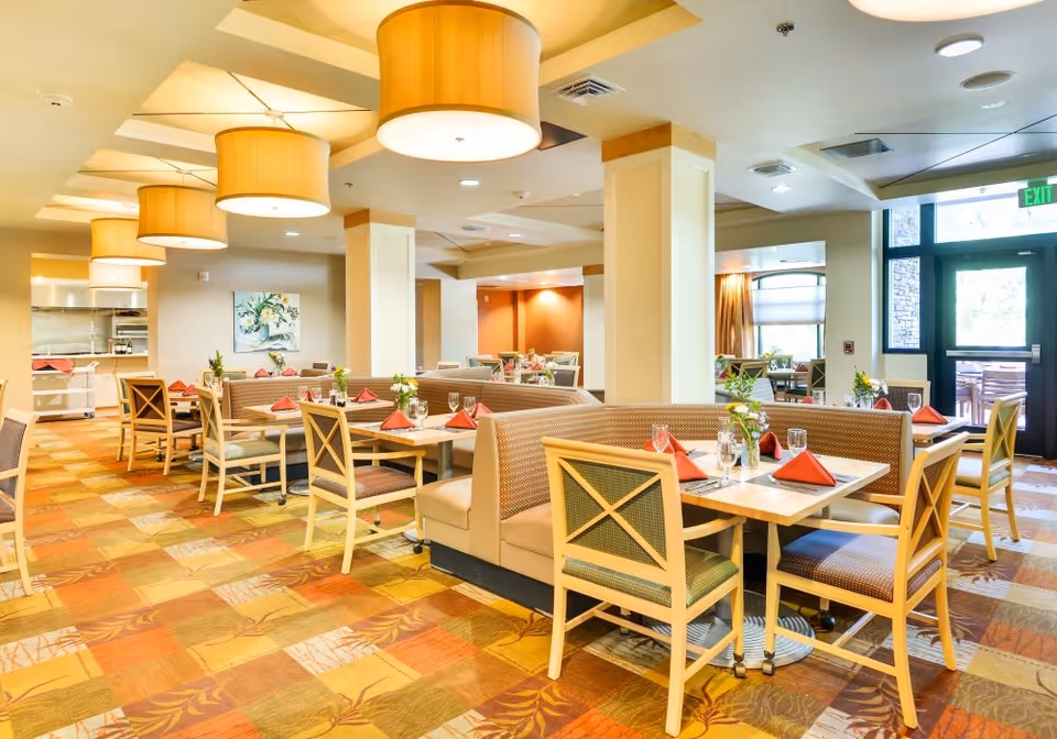 Well-lit dining room with booths and tables set with red napkins and glassware, pendant lights overhead, and a patterned carpet.