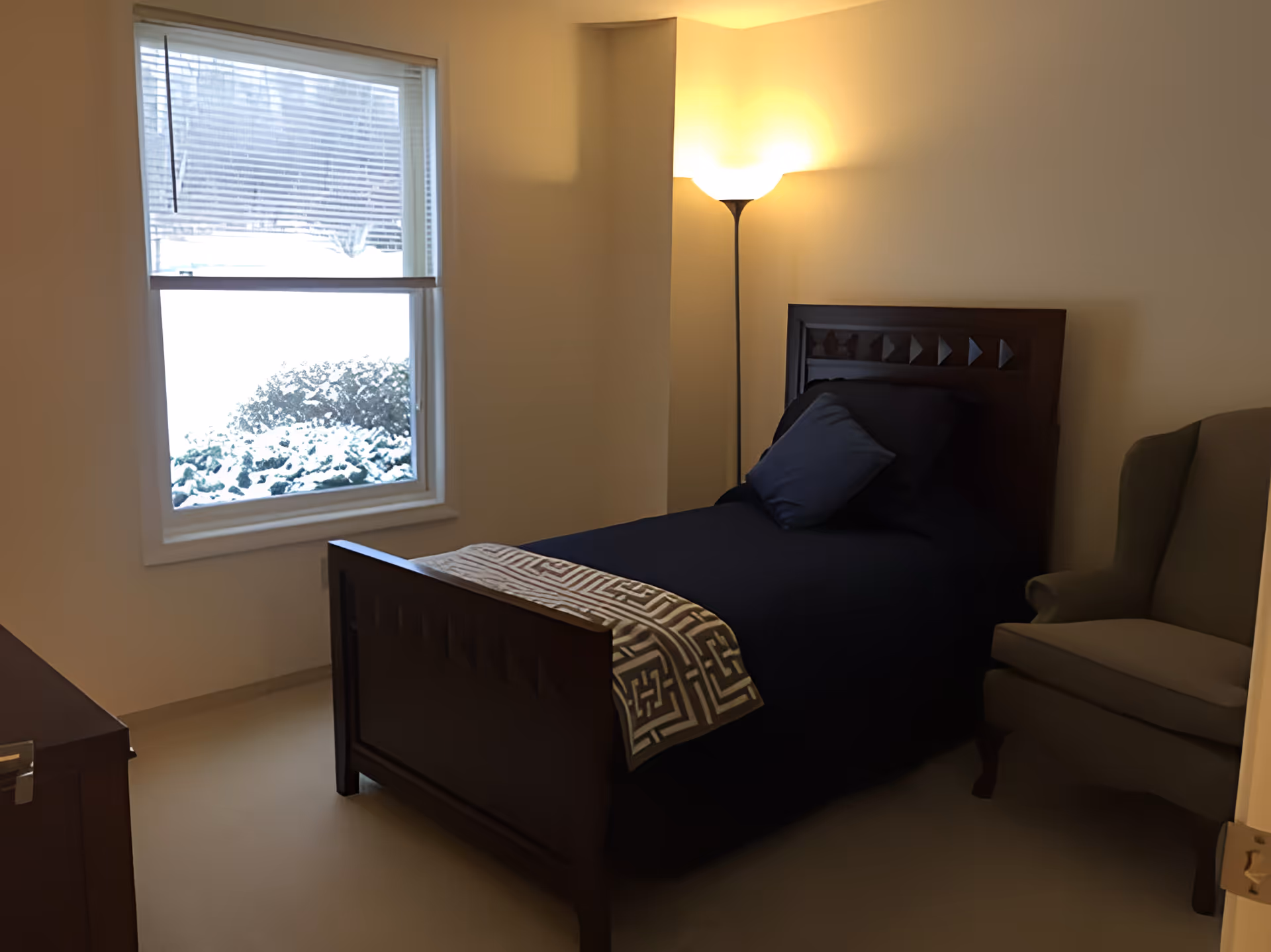 A small bedroom with a single dark wooden bed made with dark blue bedding and a patterned throw blanket at the foot. There is a matching dark blue pillow and a smaller pillow on the bed. A tall floor lamp is lit in the corner next to the bed, and a beige armchair is positioned to the right. A window on the left shows a snowy outdoor scene with bushes covered in snow.