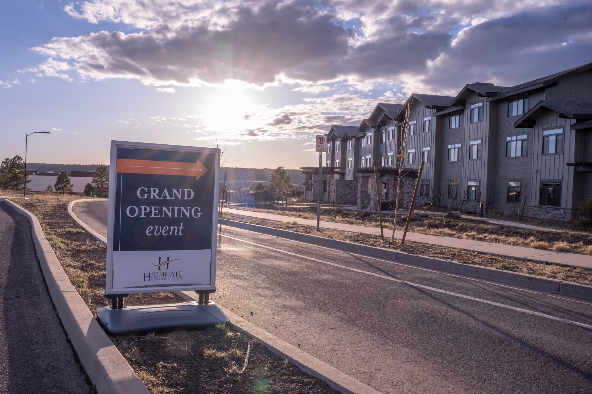 A 'Grand Opening event' sign sits by a roadway in front of a large multi-story senior living building under a cloudy sunset sky.