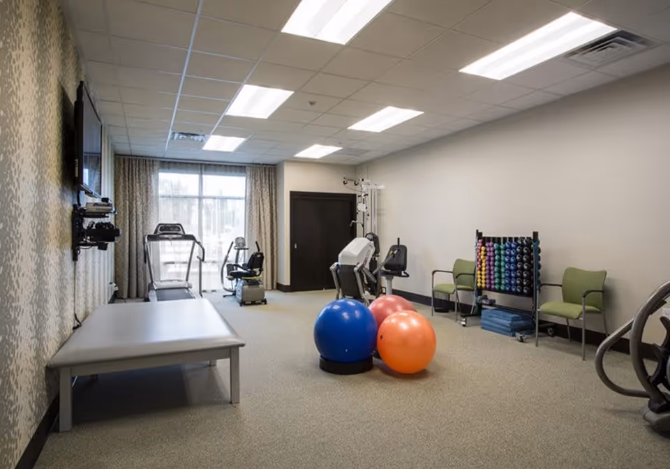 A fitness room in a senior living facility with exercise equipment including a treadmill, stationary bike, and weight machine. There are three large exercise balls in blue, pink, and orange on the carpeted floor. Two green chairs and a rack with colorful dumbbells and exercise mats are against the wall. A flat-screen TV is mounted on the wall, and large windows with curtains allow natural light into the room.