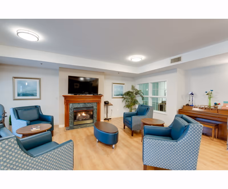 Bright common lounge with blue upholstered chairs arranged around a fireplace with a TV above, small tables, and a piano in the corner.