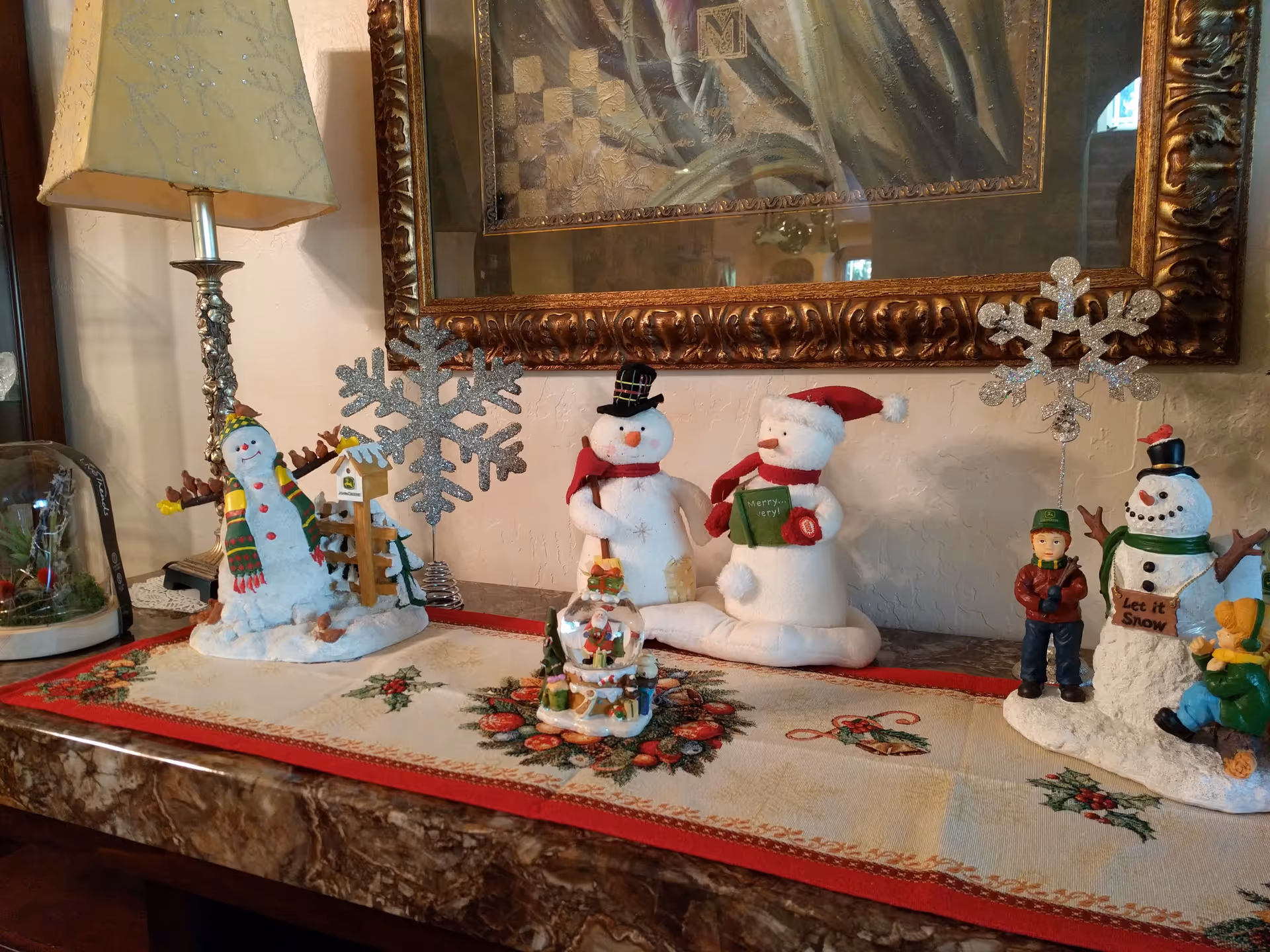 A decorative holiday display on a marble surface featuring various snowman figurines, a snow globe with Santa inside, and glittery snowflake ornaments. The display is set on a festive table runner with holly and berry patterns, beneath a large ornate framed painting and next to a vintage-style lamp.