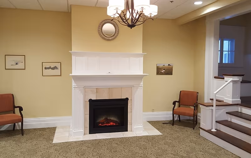 Small common room with a white fireplace and electric fire, two upholstered chairs, wall art, chandelier and a short staircase.