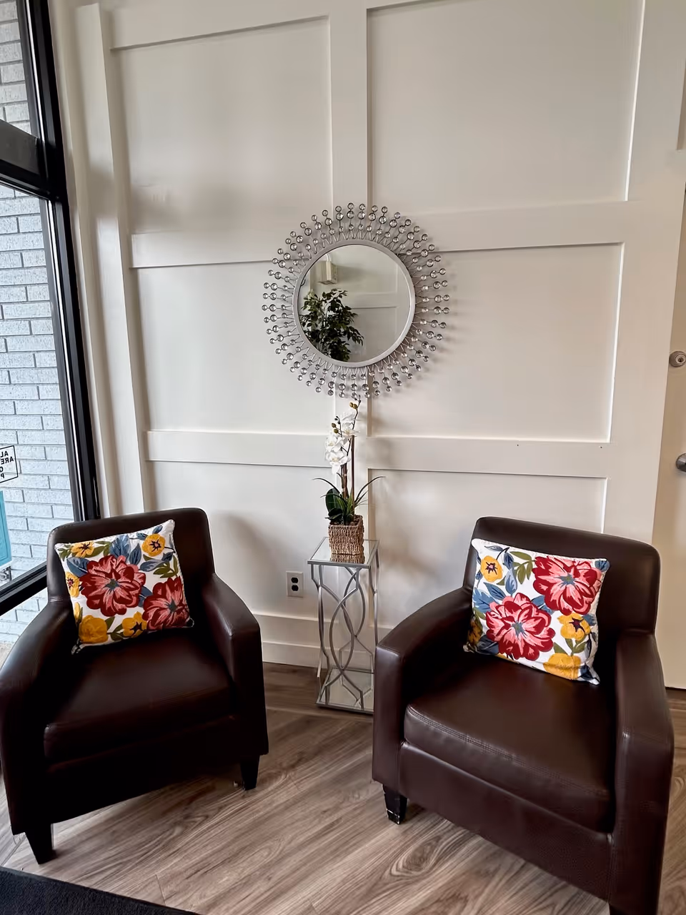 Two brown armchairs with colorful floral pillows flank a small glass side table topped with a potted orchid beneath a decorative round mirror on a white paneled wall.