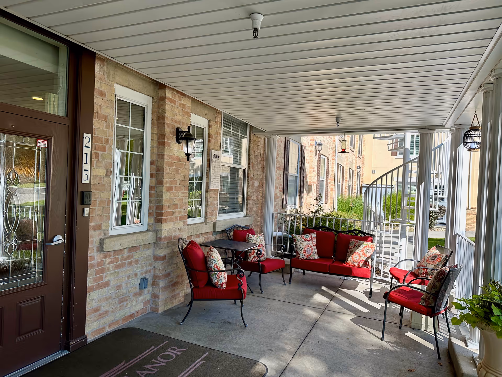 Covered outdoor patio area with red cushioned chairs and a loveseat arranged around a small table. The patio has a concrete floor, brick walls with windows, a brown door with the number 215, and a spiral staircase leading upwards. There are decorative pillows on the chairs and some potted plants visible.