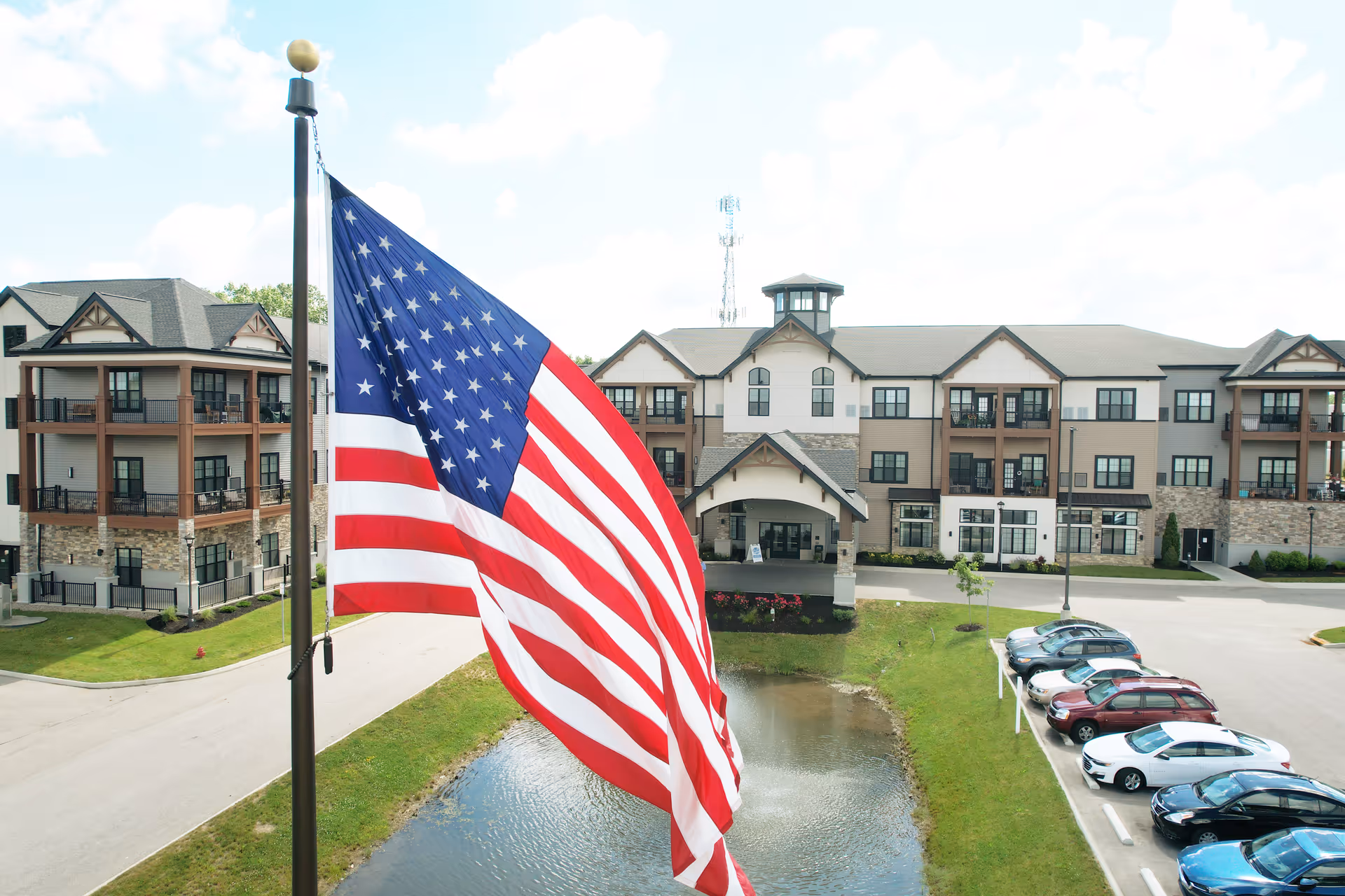View of a senior living facility building with multiple balconies, a pond in front, a parking lot with cars, and a large American flag on a flagpole in the foreground.