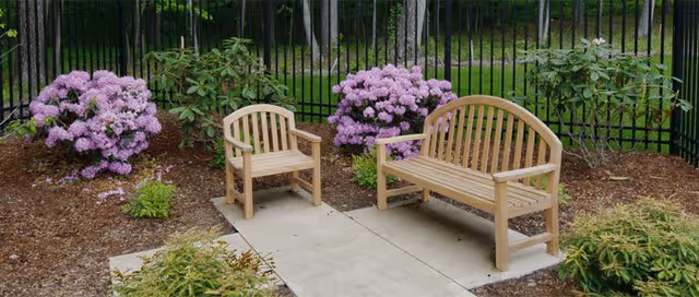 Wooden bench and chair on a small paved patio surrounded by blooming purple rhododendron bushes and a black metal fence.