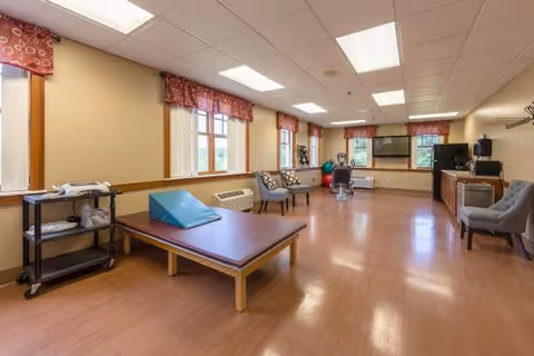 Bright, spacious rehabilitation room with a therapy table, chairs, and exercise equipment and windows along one wall.