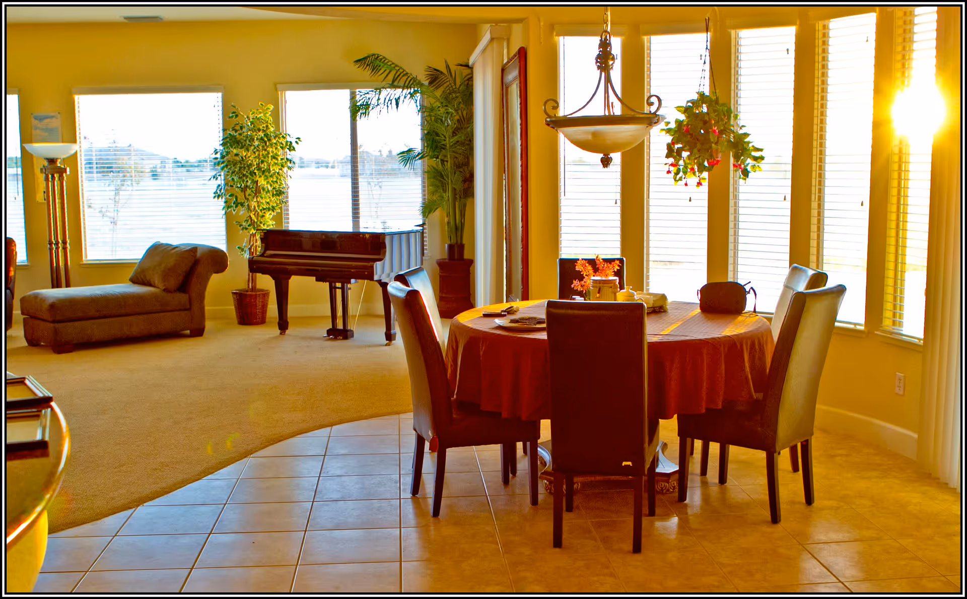 A bright and spacious living and dining area with large windows letting in natural light. The dining area features a round table covered with a tablecloth and surrounded by six chairs. A hanging plant and a decorative light fixture are above the table. The living area has a chaise lounge, a floor lamp, potted plants, and a grand piano near the windows.