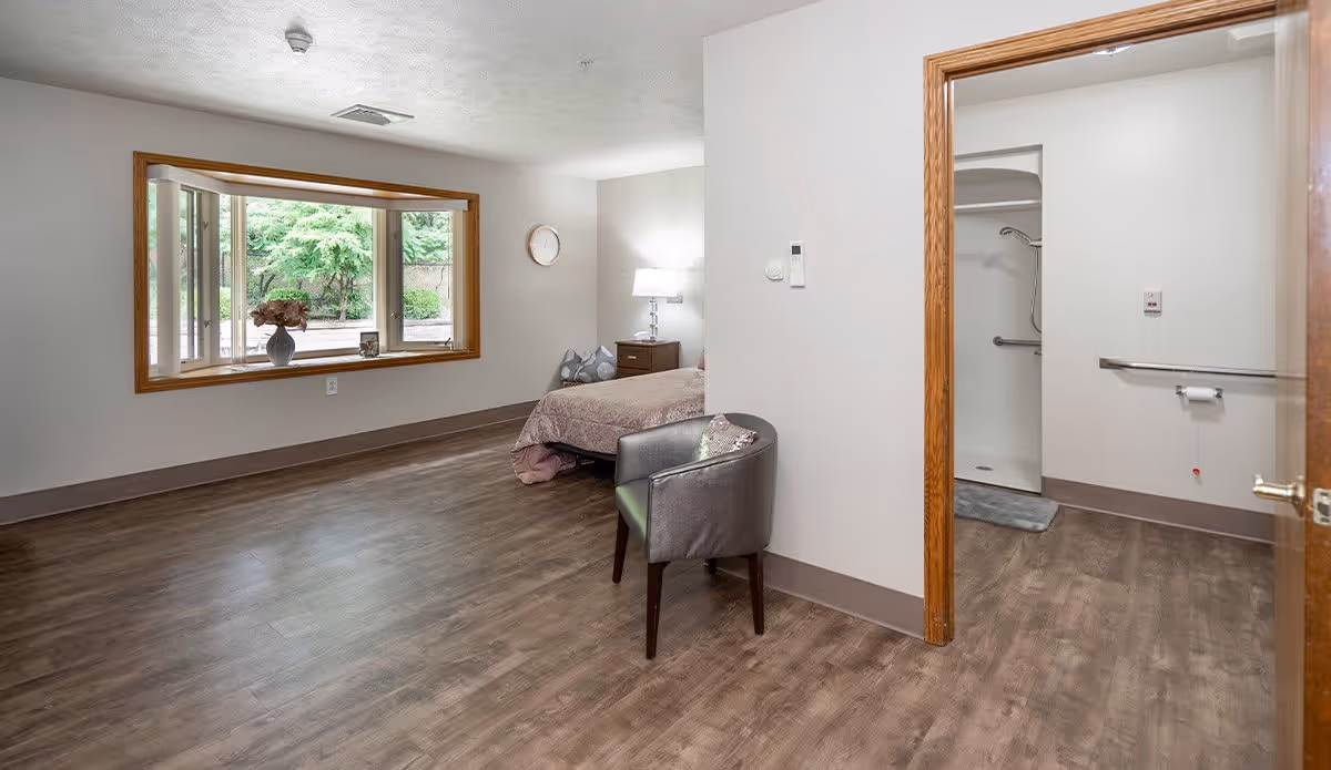 A senior living facility room with a bed, a bedside table with a lamp, a gray armchair, and a large window with a view of greenery outside. To the right, there is an open doorway leading to an accessible bathroom with grab bars and a shower area.