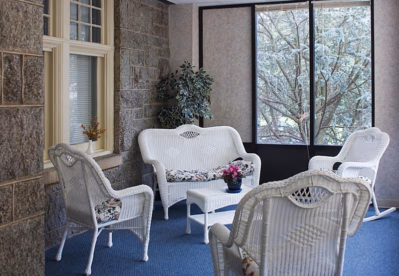 A cozy indoor seating area with white wicker furniture including a loveseat, three chairs, and a small table with a potted plant. The room has large windows showing trees outside and stone walls with a window on one side.