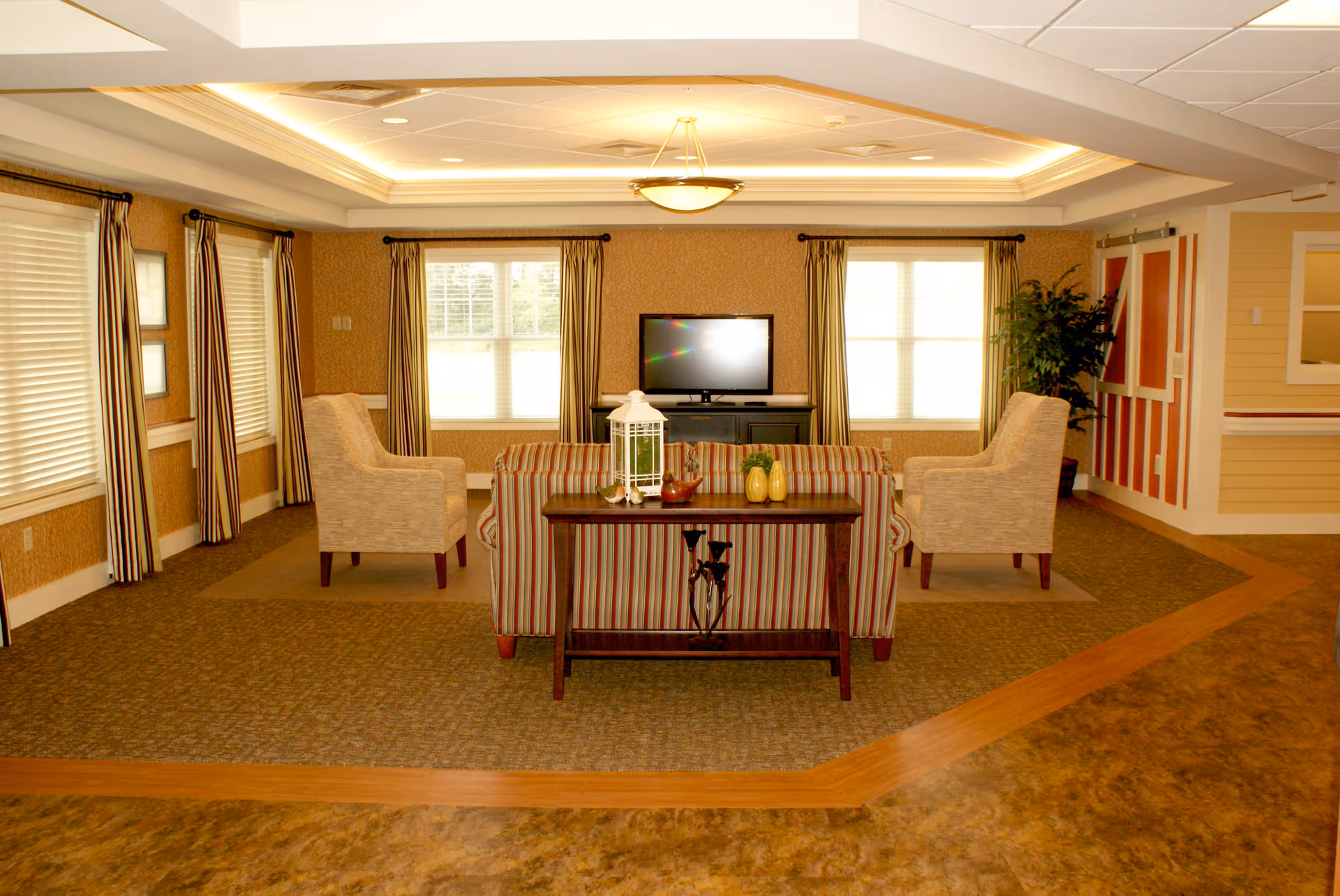 A well-lit communal living room with a striped sofa and two armchairs arranged around a coffee table facing a television between two windows.