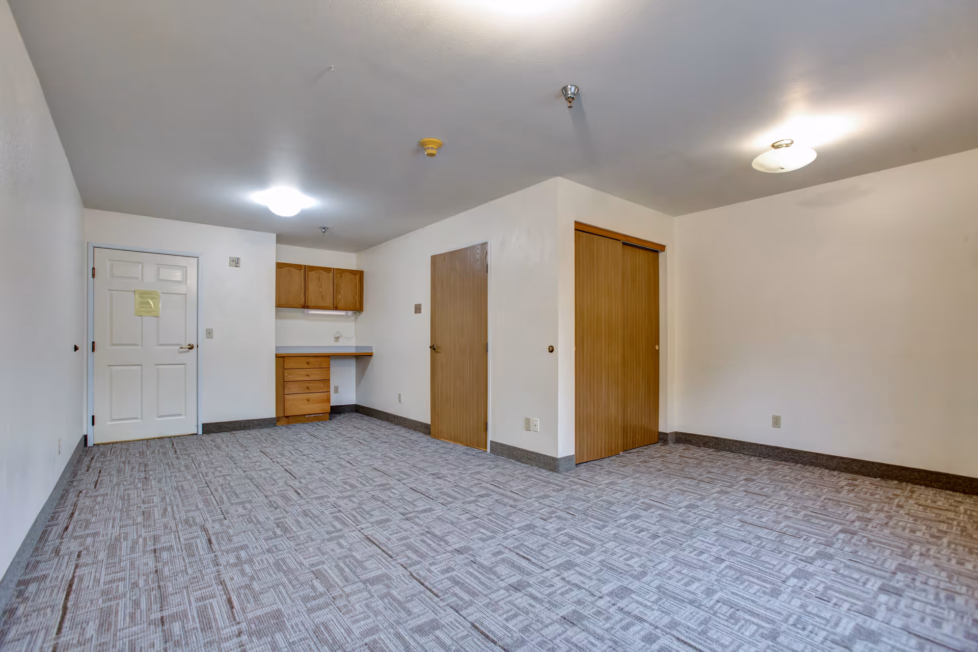 Empty room with light gray patterned carpet, white walls, and ceiling lights. There is a white door on the left wall, a small built-in wooden desk with cabinets above it, a wooden door in the center, and a wooden sliding closet door on the right wall.