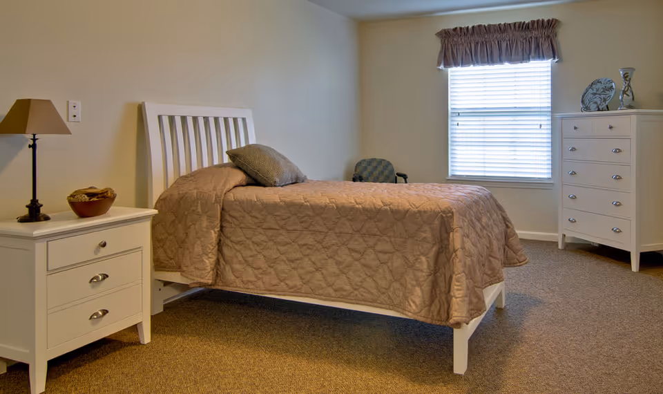 Furnished bedroom with a single bed, nightstand and lamp, dresser, chair, and a window with blinds.