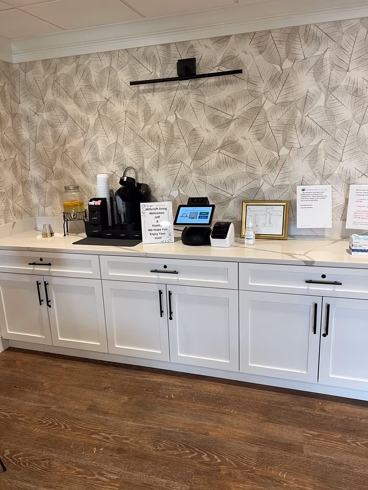 A countertop area in Millcroft Living with white cabinets and a marble-like surface. On the counter, there is a beverage dispenser with a yellow drink, a coffee machine with cups and condiments, a digital check-in kiosk, hand sanitizer, and several framed notices and signs. The wall behind has a leaf-patterned wallpaper and a black picture light above.