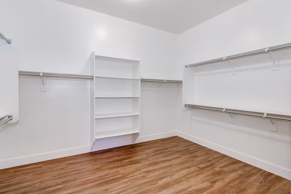 Empty walk-in closet with white walls, multiple metal hanging rods, and built-in white shelving unit. The floor is wood with a warm brown finish.
