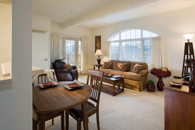 A cozy living room and dining area with a wooden dining table set for four, a brown sofa with patterned cushions, a leather armchair, a coffee table, a side table with a lamp, large arched window with curtains, and decorative plants and flowers.
