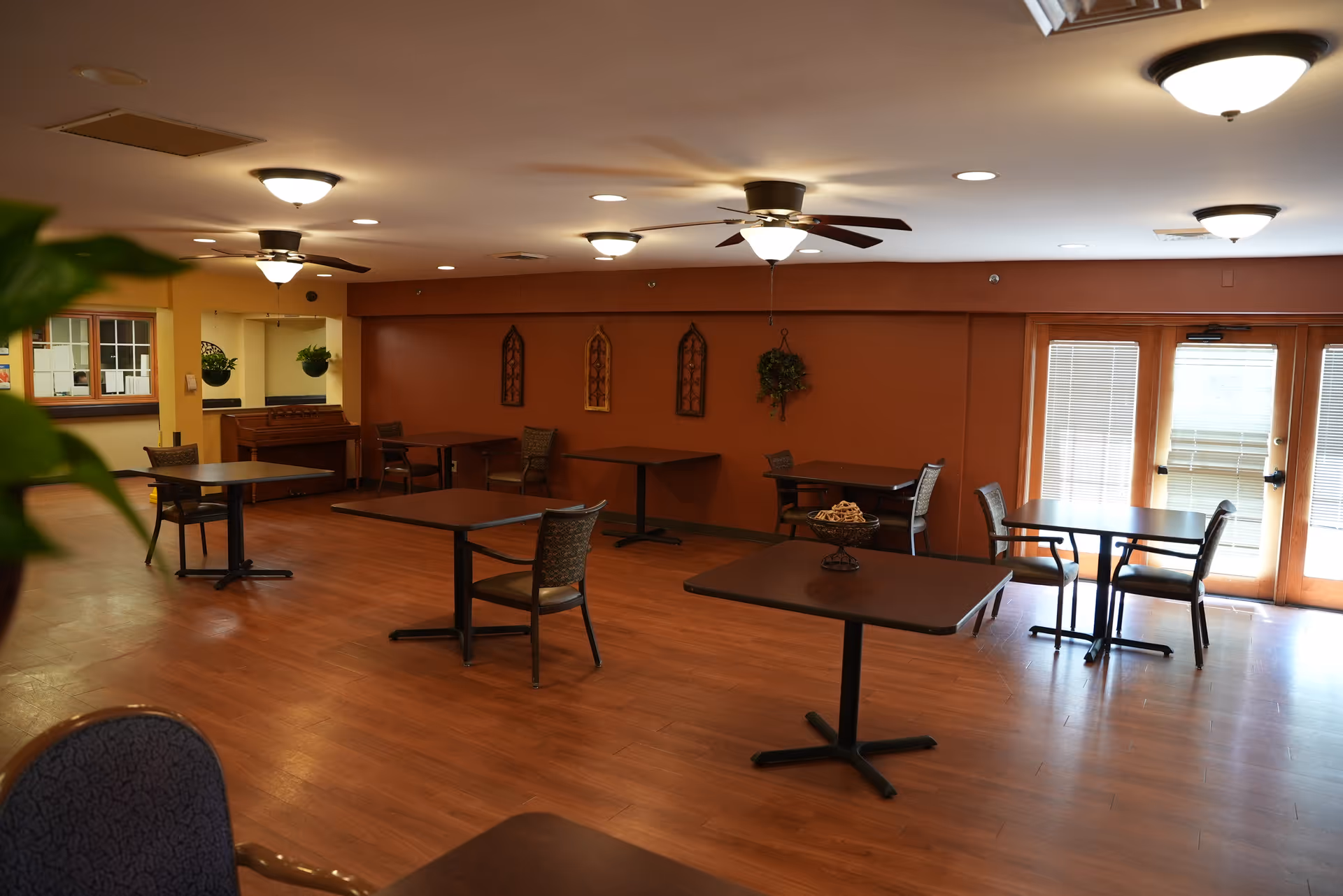A spacious dining room with several square tables and chairs arranged throughout. The room has wooden flooring, ceiling fans with lights, and large windows with blinds allowing natural light to enter. There are decorative wall hangings and plants adding to the cozy atmosphere.