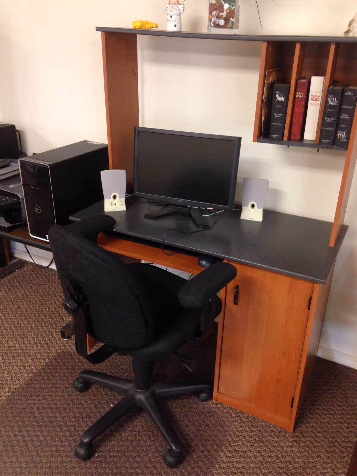 A computer workstation with a black office chair, a desktop computer with monitor and speakers on a wooden desk. There are several books, including multiple Bibles, on a shelf above the desk. The floor is carpeted.