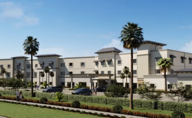 Exterior view of a large, modern senior living facility building with three stories, surrounded by palm trees, shrubs, and a well-maintained lawn. Several cars are parked near the entrance, and a few people are walking nearby under a clear blue sky.