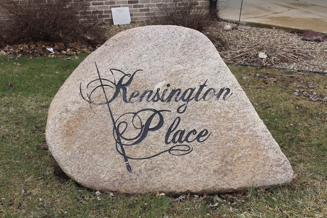 A large natural stone with the words 'Kensington Place' artistically painted on it, placed on a grassy area with some dry leaves and a brick building wall in the background.